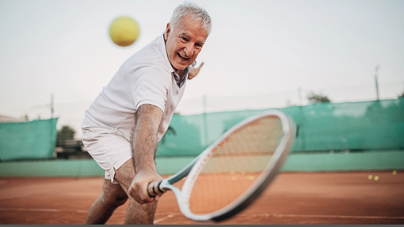 Eine ältere Frau mit Tennisschläger und Ball beim Aufschlag.