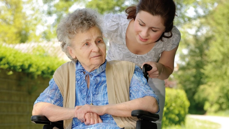 Junge Frau spaziert mit Seniorin im Rollstuhl.
