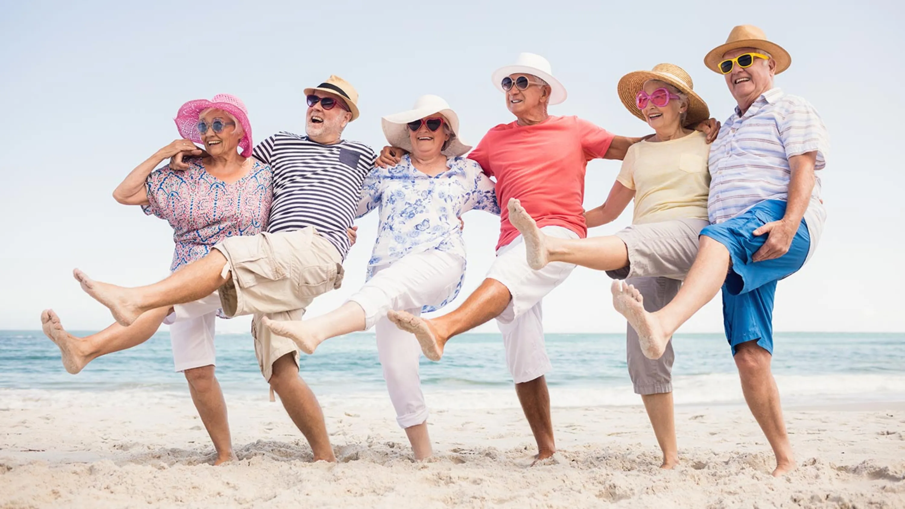Sechs fröhliche ältere Frauen und Männer mit Hüten und Sonnenbrillen stehen in einer Reihe am Strand und heben tanzend und lachend ein Bein.